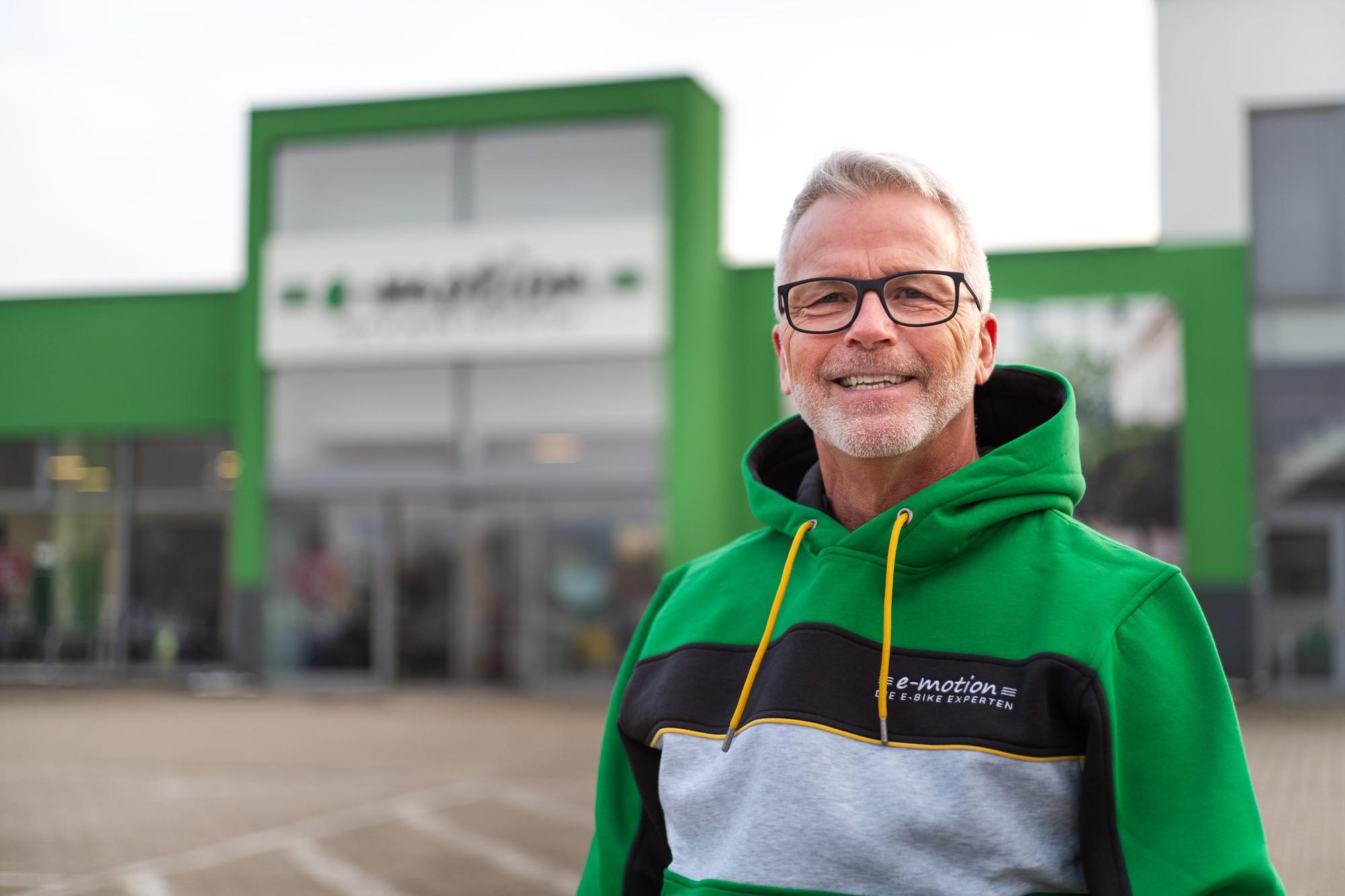 Auf dem Bild steht ein lächelnder Mann im Vordergrund, der als Shopleiter eines E-Bike-Geschäfts fungiert.