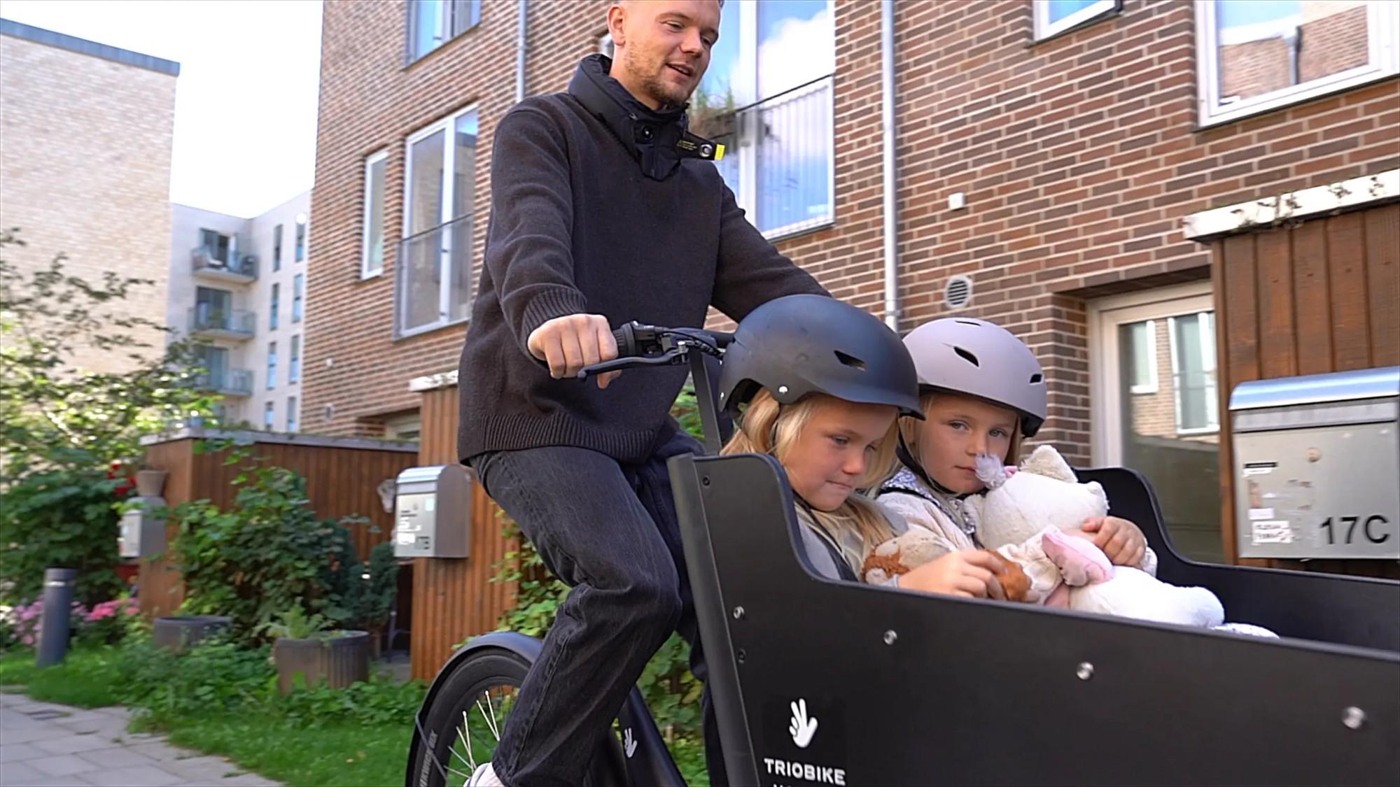 Ein Mann fährt mit dem Triobike Hafnia Lastenrad und zwei Kindern in der Transportbox durch die Stadt