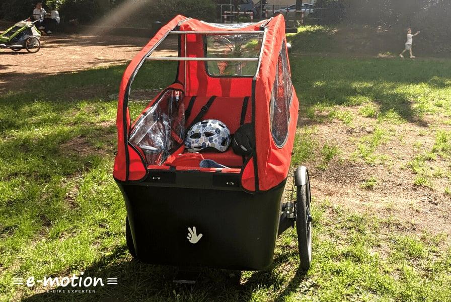 Roter Fahrradanhänger im Park mit Fußball und Kinderwagen im Hintergrund