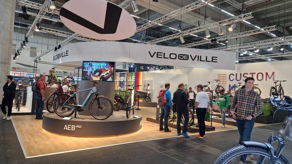 Moderner Messestand mit e-Bikes von Velo de Ville und AEB Pro, Zubehör und interessierten Besuchern.
