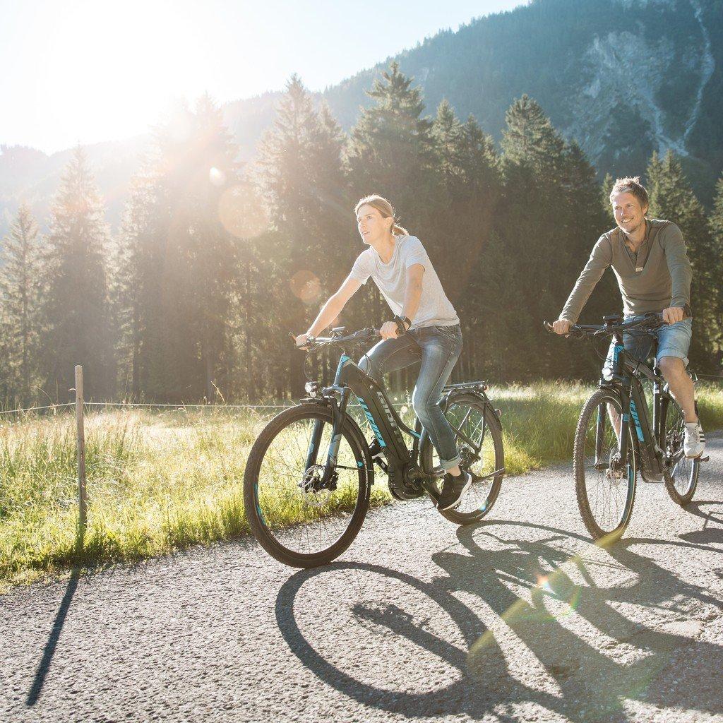Zwei Personen fahren lächelnd auf e-Bikes auf einem sonnigen Weg durch eine malerische Landschaft mit Wald im Hinte