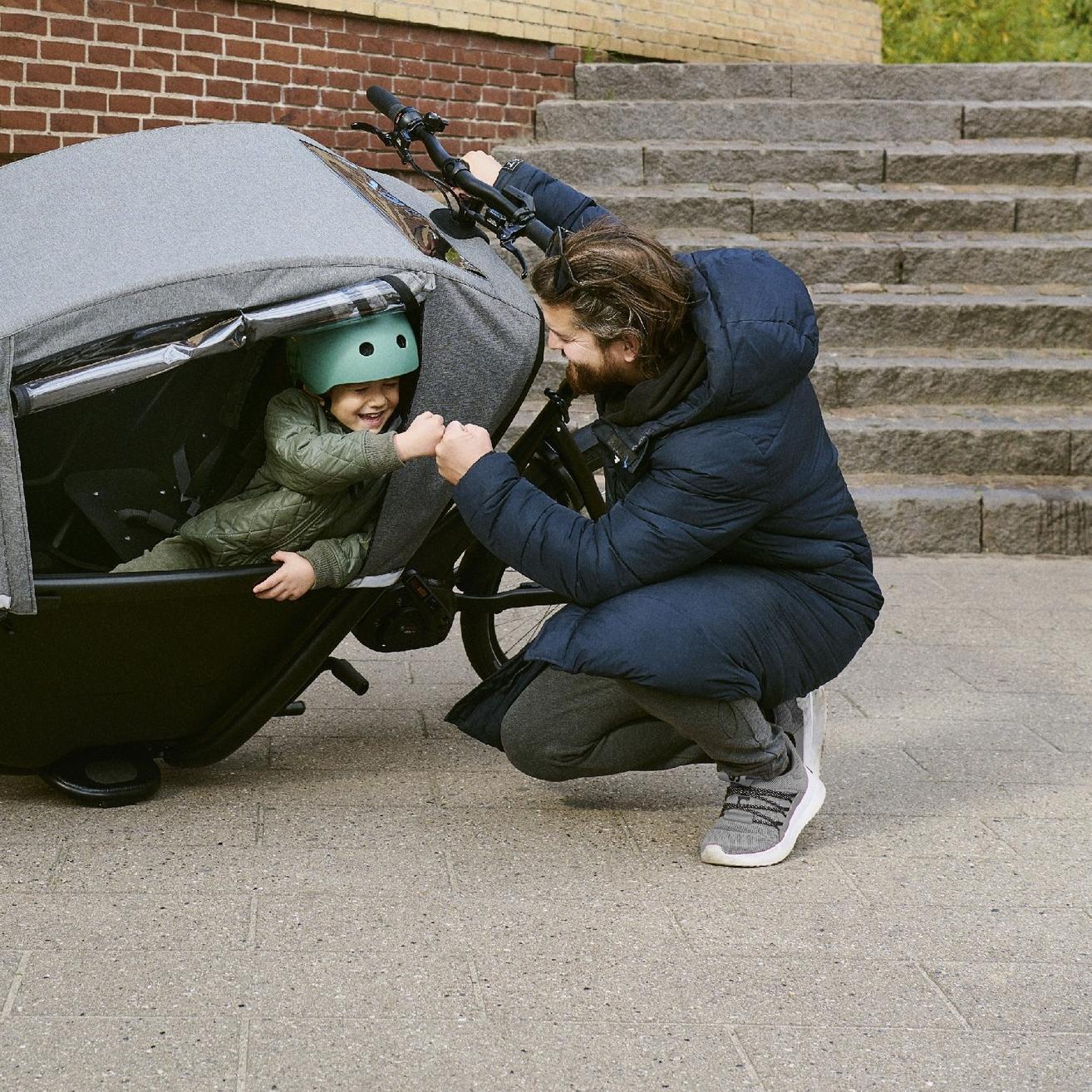 Ein Mann hockt neben einem Winther Lastenrad und schlägt bei einem Kind ein, welches in der Transportbox sitzt