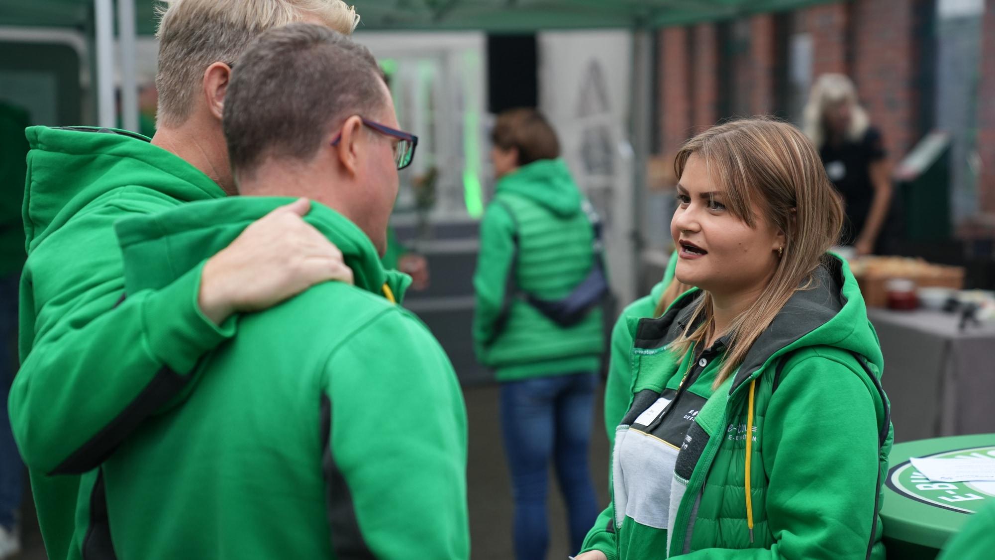 Drei Personen in grüner Kleidung unterhalten sich bei einem Event. Markenlogos sind auf der Kleidung sichtbar.