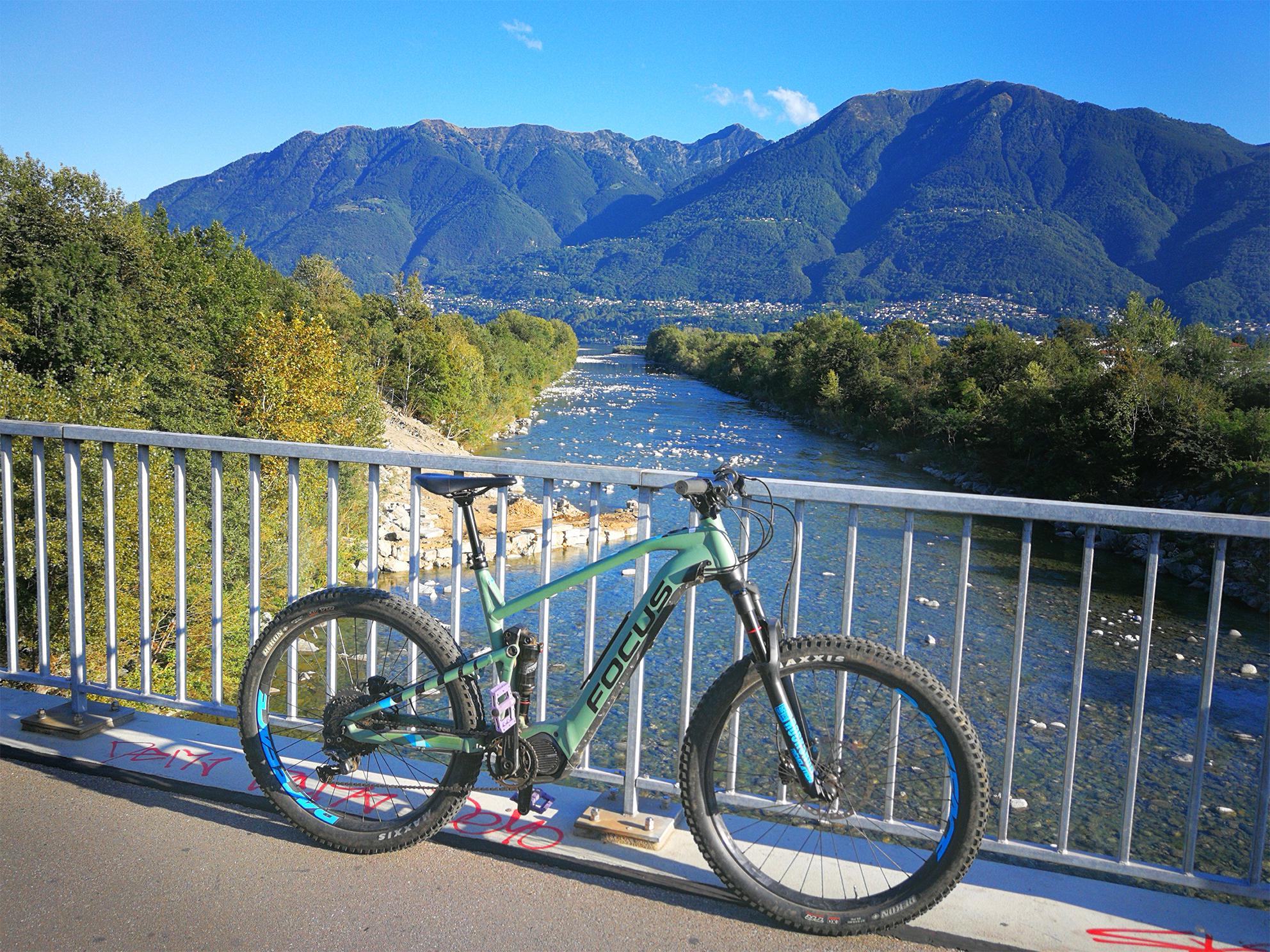 Urlaubsbild mit e-Bike, es lehnt an einem Geländer, im Hintergrund Wasser und Berge