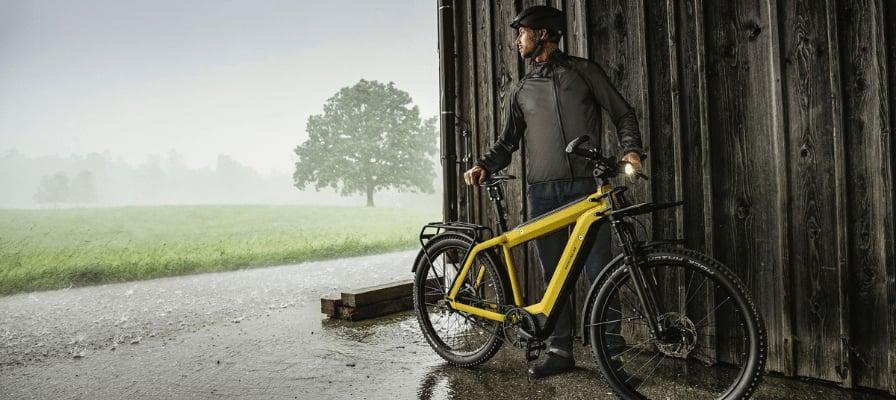 Mann mit gelbem E-Bike steht bei Regen vor Holzhütte