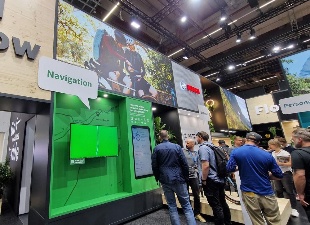 Messestand zu e-Bike-Technologie, Besucher am interaktiven Bildschirm mit Fokus auf Navigation.