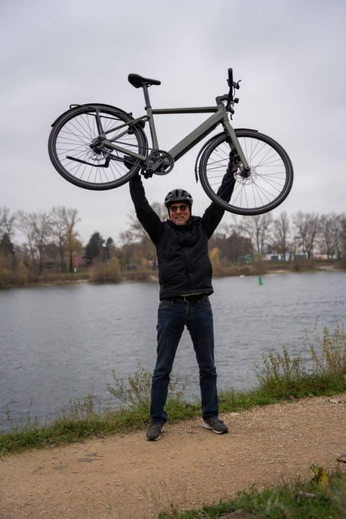 Mann hebt E-Bike vor Flusslandschaft in die Luft