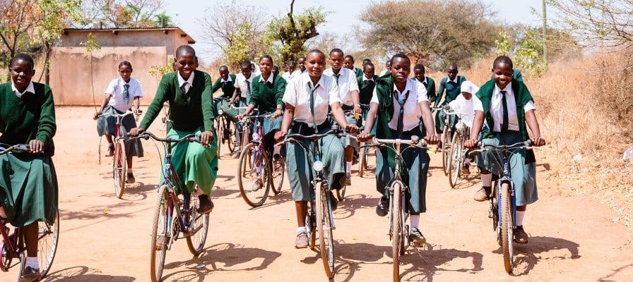 Schülerinnen fahren gemeinsam mit Fahrrädern zur Schule auf einem staubigen Weg