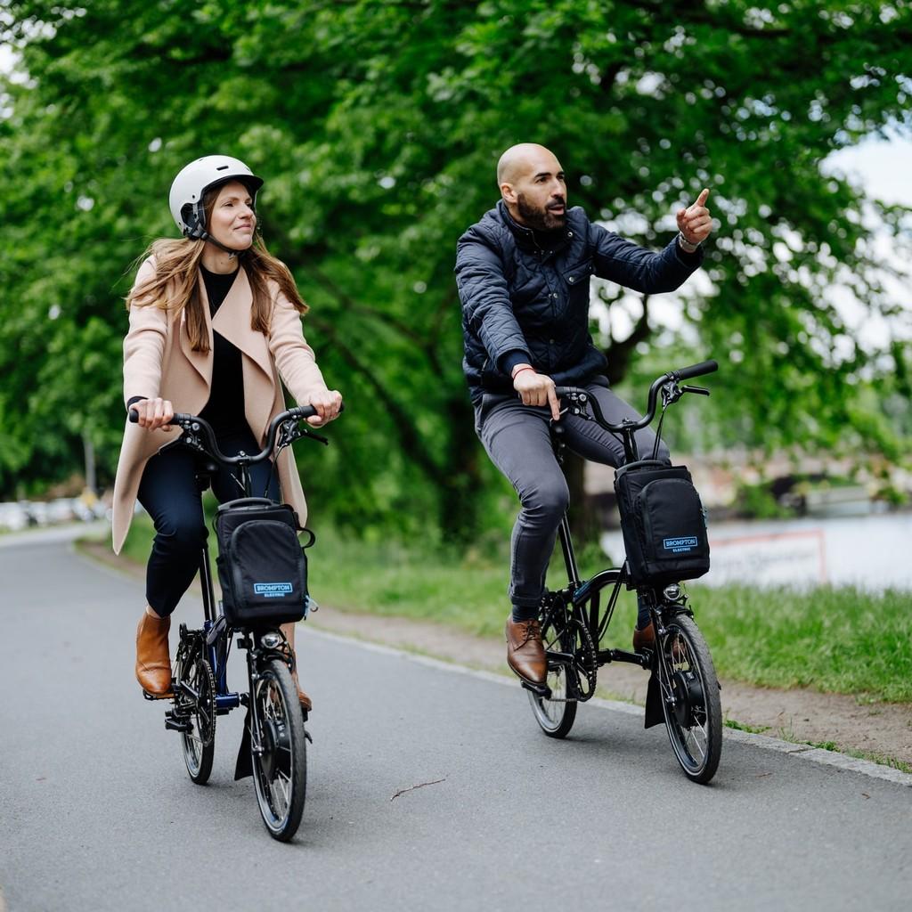 Zwei Personen fahren Brompton-Falträder entlang eines malerischen Weges. Ideal für urbane Mobilität und stilvolle A