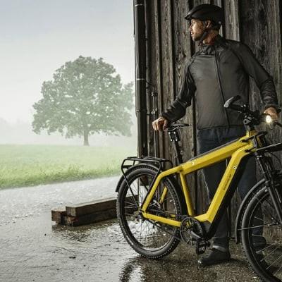 Mann mit gelbem E-Bike steht bei Regen vor Holzhütte