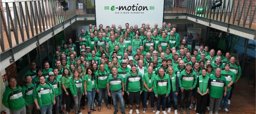 Große Gruppe von Personen in grüner Kleidung vor e-motion Logo. E-Bike Enthusiasten und Experten vereint.