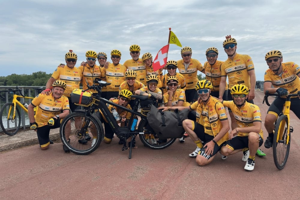 Gruppe von Radfahrern in gelben Trikots mit E-Bike und Flaggen im Freien