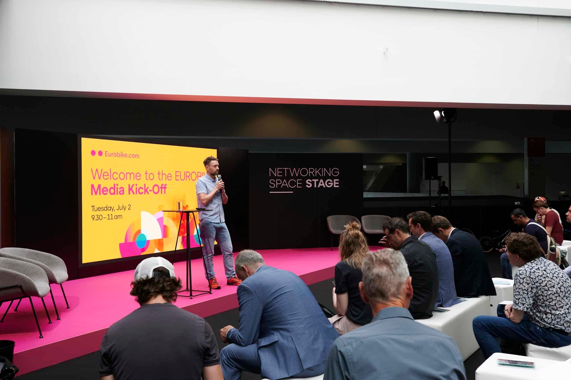 Präsentation auf pinkfarbener Bühne mit Redner, großem Bildschirm und aufmerksamem Publikum.