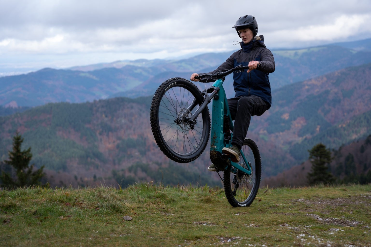 Das Bild zeigt einen Fahrer, der auf einem Focus Jam² 7.8 E-Bike ein Wheely in einer bergigen Landschaft ausführt.