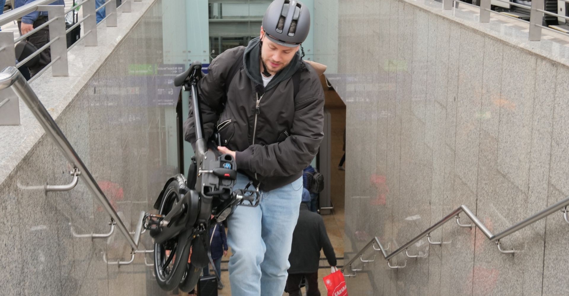 Das Bild zeigt einen Mann, der eine zusammengeklappte Brompton-Faltradtreppe in einem Bahnhof hinaufträgt.
