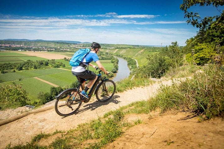 Person auf E-Bike fährt auf einem Hügel mit Flussblick bei sonnigem Wetter