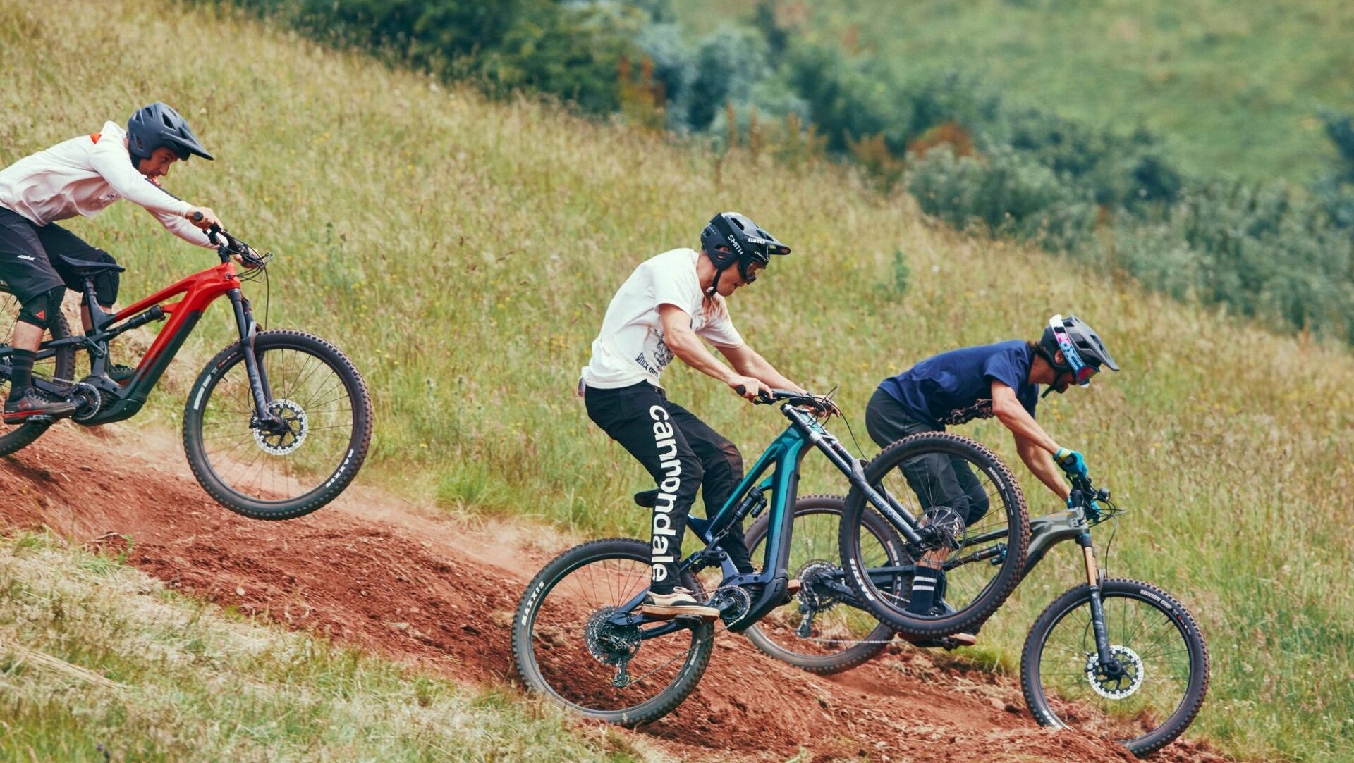 Drei Mountainbiker mit Helmen fahren Cannondale Moterra Neo e-MTBs eine staubige Abfahrt im Grünen hinunter.